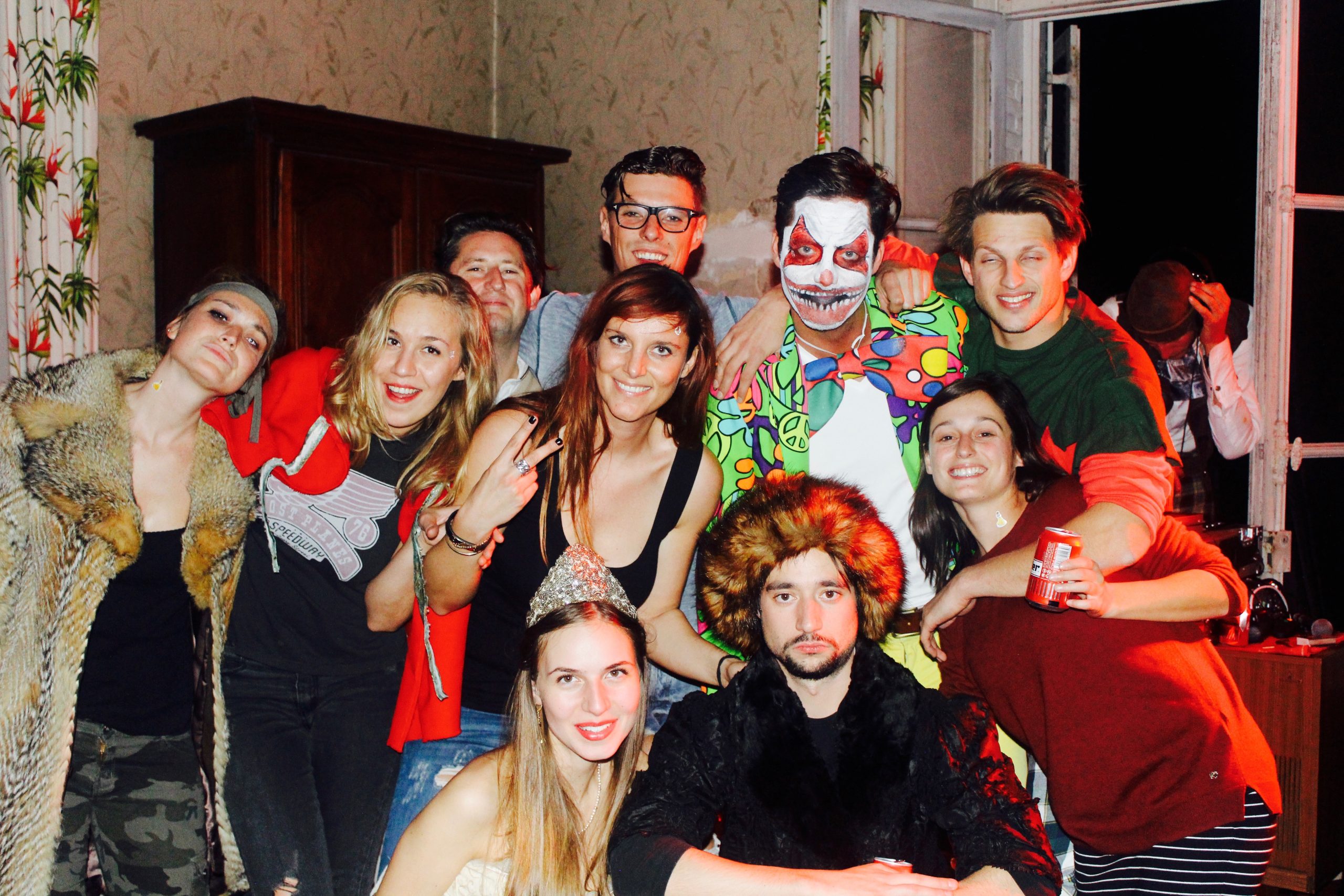 Een groep vrienden viert Halloween in een sfeervolle kasteelkamer. Ze lachen en poseren samen, sommigen in kostuum — één persoon draagt een kleurrijk clownspak met griezelige schmink, anderen hebben bontjassen, kronen of feestelijke accessoires aan. De warme verlichting en open ramen geven het moment een gezellige en spontane sfeer.