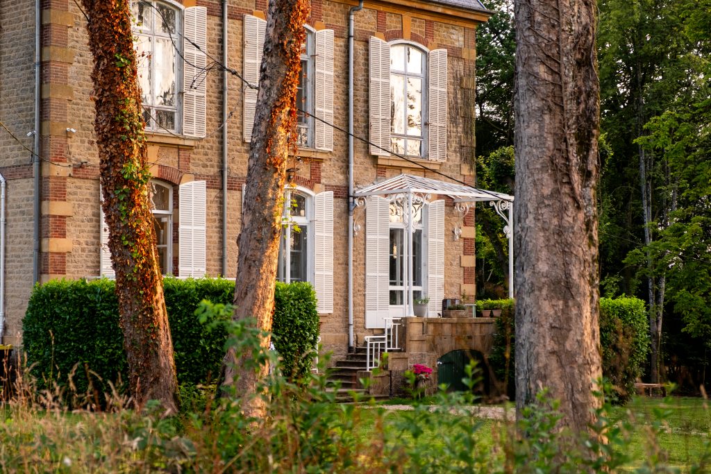 Elegant stone château façade at Château Guillermo in the French Ardennes, surrounded by tall trees and greenery in warm golden evening light.