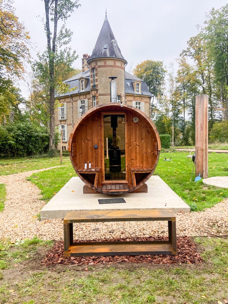 Outdoor barrel sauna in the garden of Château Guillermo with the historic stone château in the background, surrounded by trees in the French Ardennes.